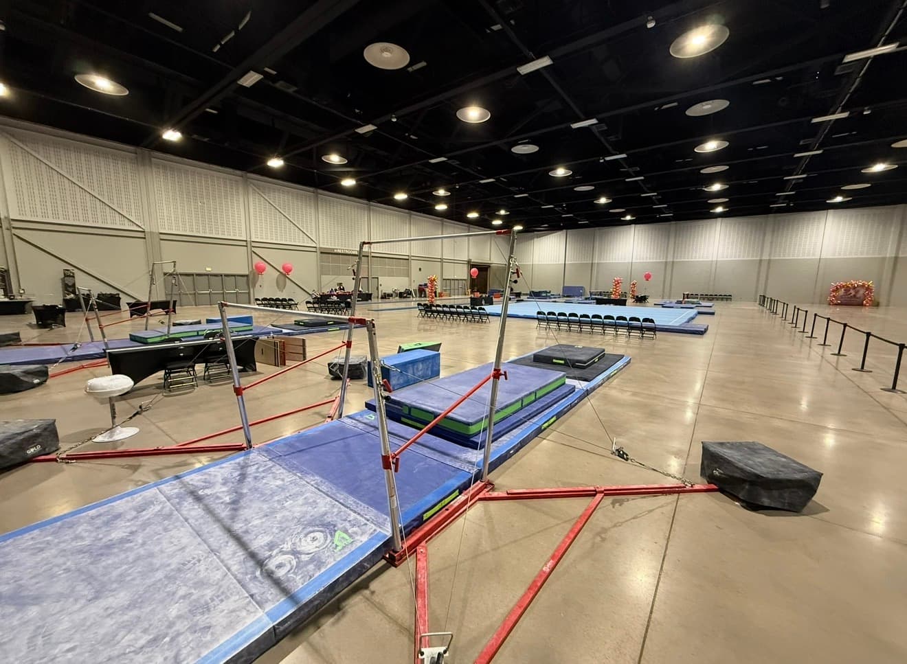 Uneven bars, mats, and a competition floor built out across a convention center hall before a meet.